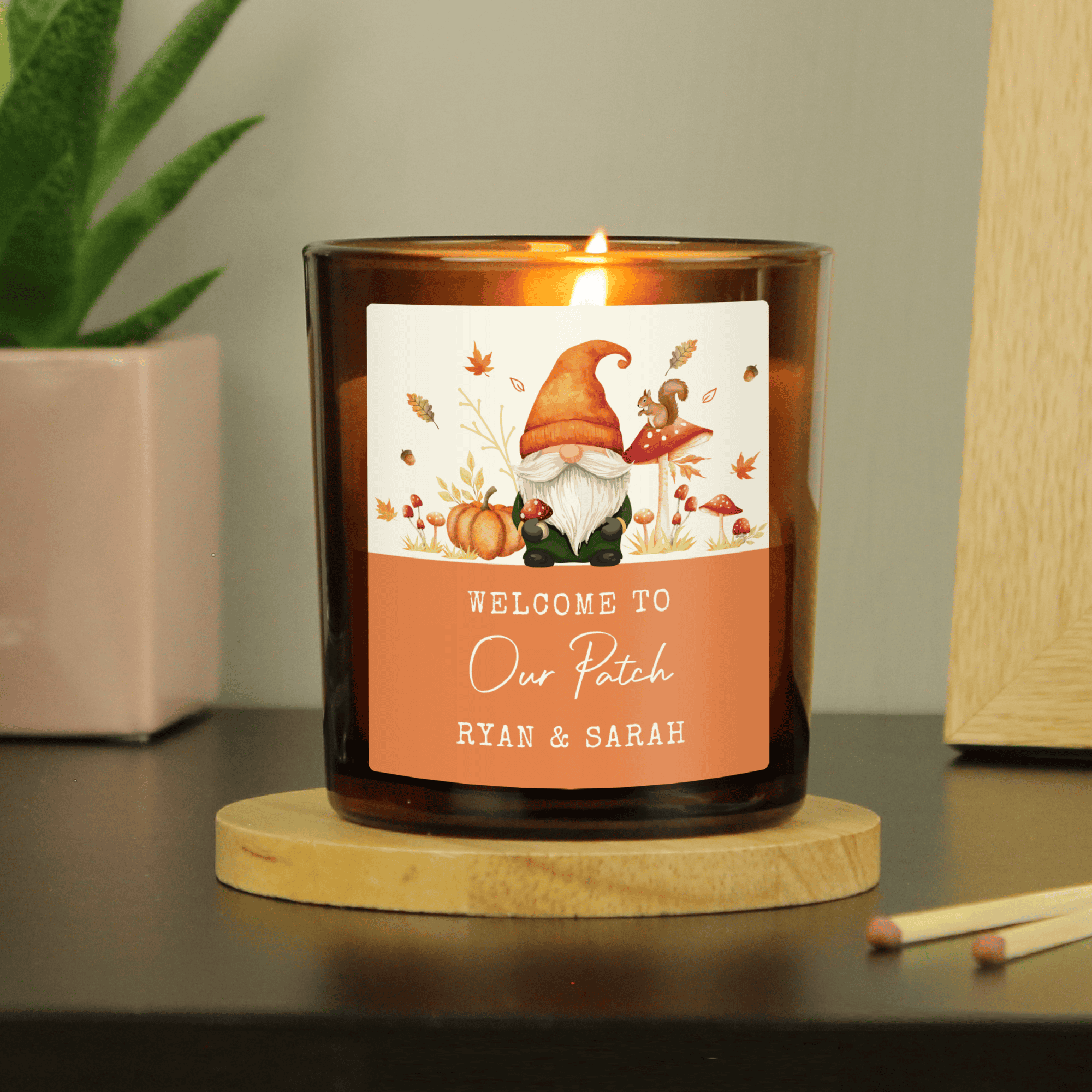 Candle with a personalised label featuring a gnome and autumn elements on a wooden coaster.